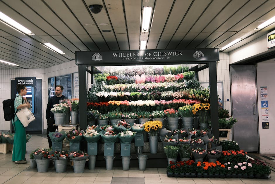 A vibrant flower display at Wheeler of Chiswick in an underground station, featuring a variety of fresh flowers arranged in metal buckets and displayed on a tiered shelving unit. The bouquet includes