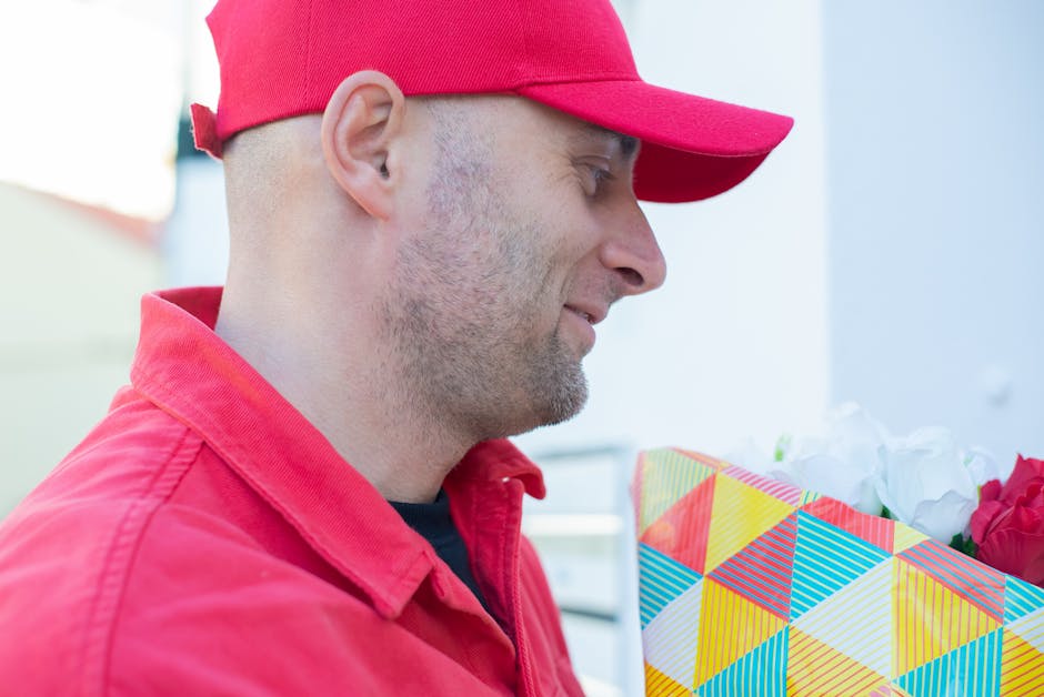 A delivery person dressed in a red cap and red uniform is holding a colorful, geometric-patterned gift bag filled with fresh red and white roses. The bouquet features vibrant open blooms with textured