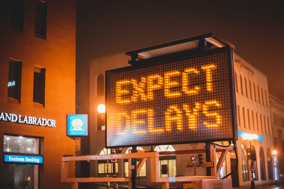 A large illuminated digital signboard on a city street at night displays the message 'EXPECT DELAYS' in bright orange letters. The sign is mounted on a metal frame with a wooden platform and is positi