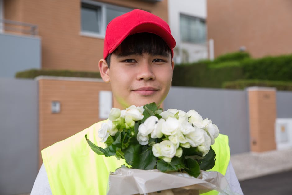 A young male flower delivery courier with light skin, dark hair, and a friendly expression, wearing a bright yellow high-visibility vest and a red cap, is holding a bouquet of fresh, white ranunculus 