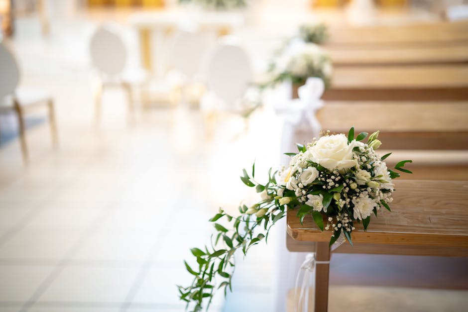 A bouquet arrangement of fresh white roses, lisianthus, and baby's breath with lush green foliage, placed on a wooden pew in a softly lit indoor setting. The bouquet is wrapped with white ribbon, and 